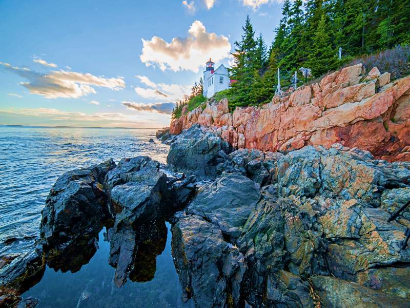 White lighthouse in the side of the oceans surrounded with the cliff of rocks and trees oin Acadia National Park