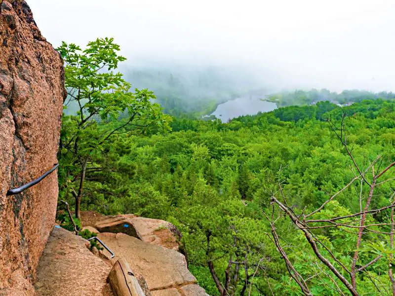 Rocks with iron bar for hiking in Acadia National Park surrounded by trees and a lake in the distance