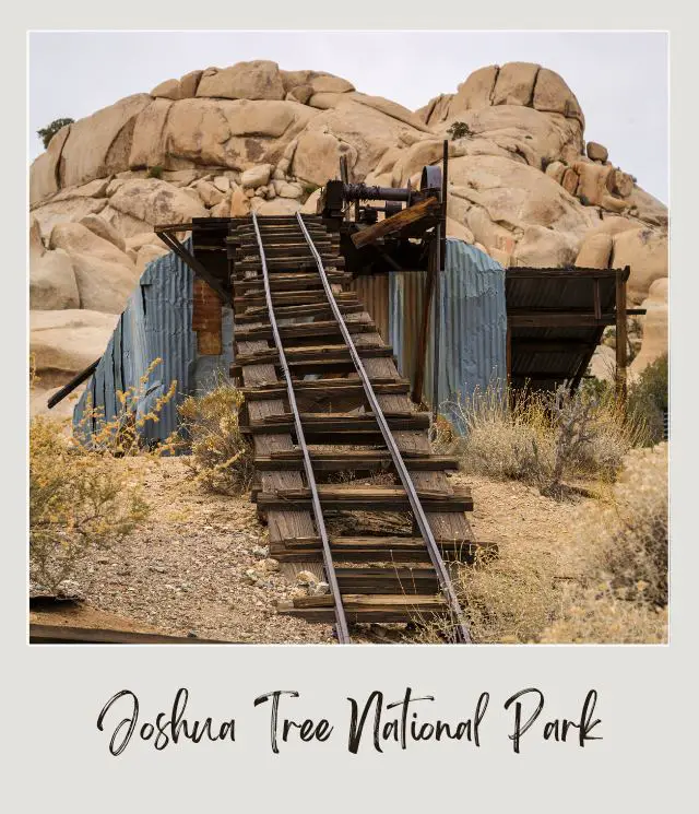 The old structure of an abandoned mine is surrounded by huge rocks, bushes, and other plants in Joshua Tree National Park.