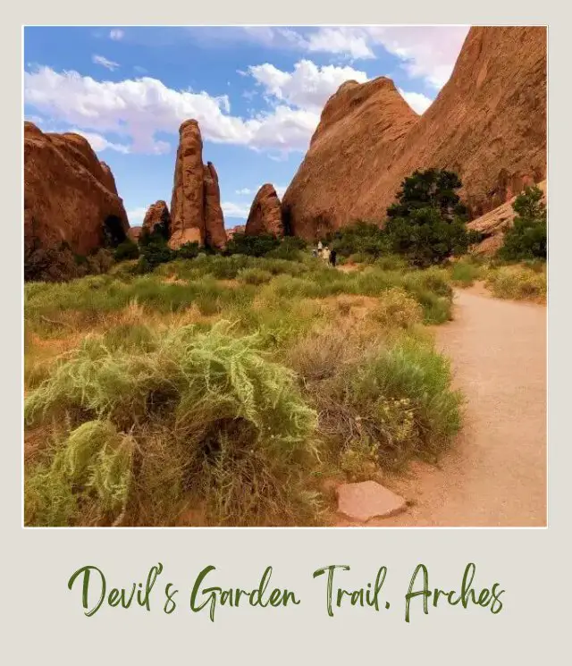 View of people walking on Devils Garden Trail surrounded by huge rock formations and bushes in Arches National Park.