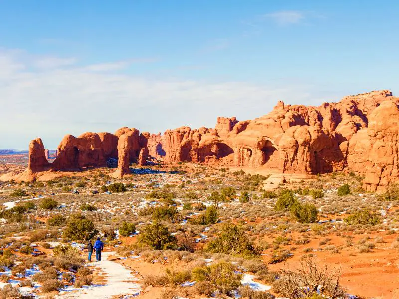 Two people surrounded by bushes covered with little snow and behind are huge rock formations in Arches National Park