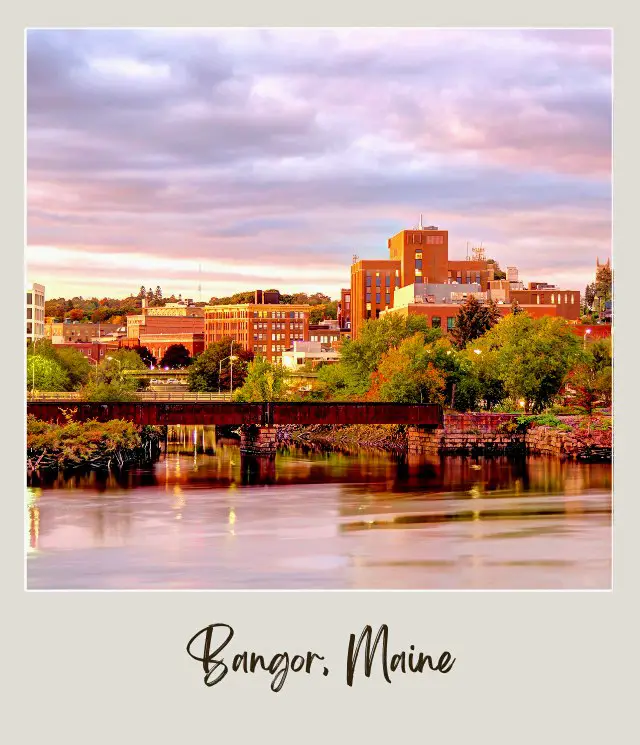 buildings with green church steeple and river in foreground with rusty bridge in Bangor Maine