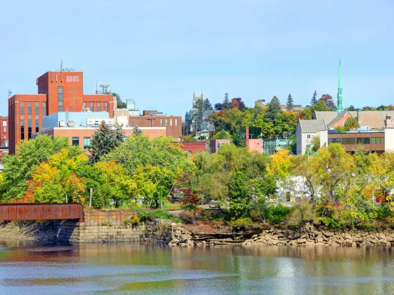 buildings with green church steeple and river in foreground with rusty bridge in Bangor Maine
