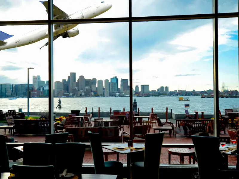 restaurant tables with large windows and plane flying close and across harbor is skyline of boston seen from boston airport