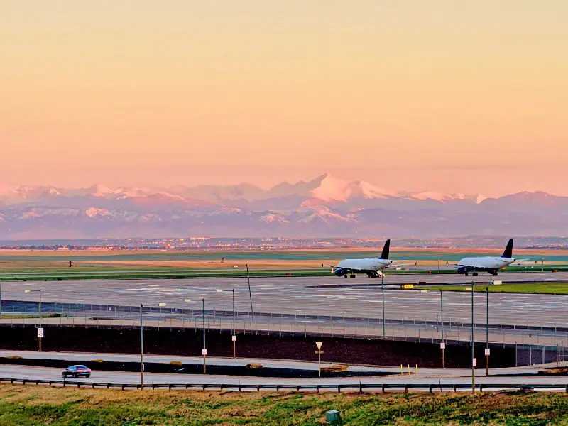 Two parked airplanes parallel each other in the middle of Denver International Airport and the mountains behind the airport.