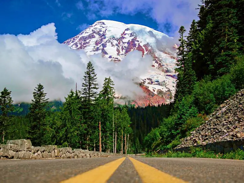 Straight road with two yellow lines in the middle surrounded by rocks and trees, and behind is the snow-capped mountain in Mount Rainier National Park
