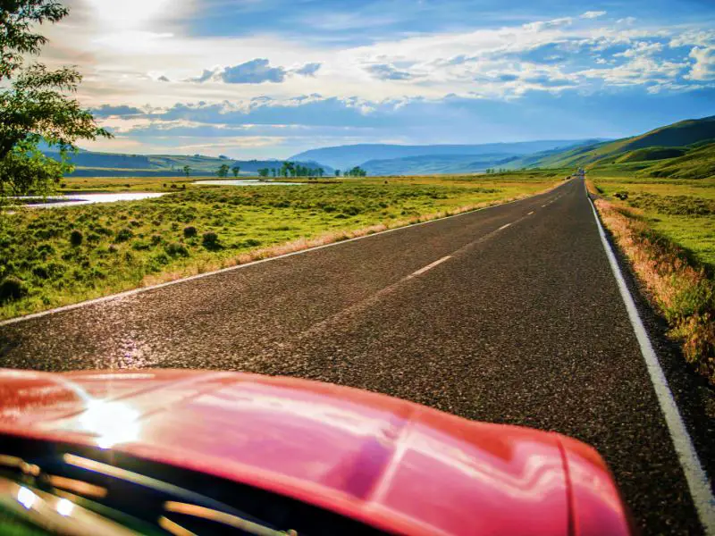 A photo of a red car hood in the middle of the straight road surrounded by green grasses and behind are mountains near Yellowstone National Park