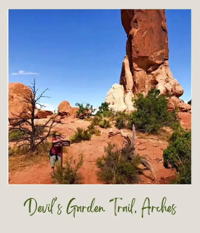 Kevin holding a wooden signage surrounded by huge rocks and bushes on Devils Garden Trail in Arches National Park.