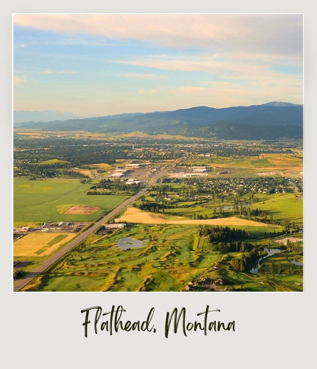 Aerial view of buildings surrounded by trees, plain fields and mountains in Flathead Country Near Glacier Park International Airport.