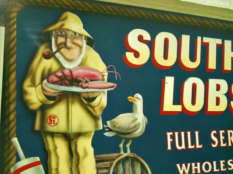 The image of a cartoon old man with a white beard holding a huge plate of Lobster and besides is a white bird in Foodie Lobster Shack, one of the things do near Acadia National Park