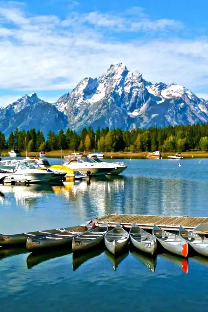 Small boats and kayaks beside the wooden bridge on the water, and behind are trees and snow-capped mountains in Grand Teton National Park.
