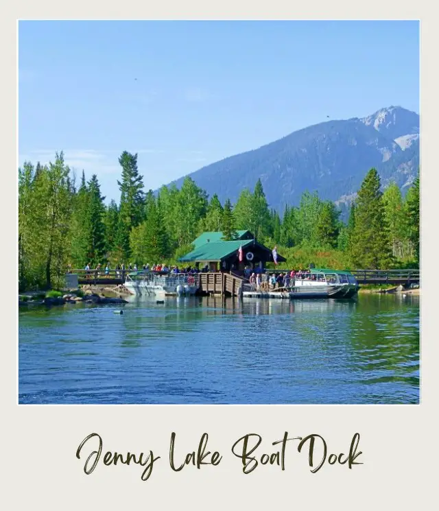 A boat dock in Jenny lake, surrounded by trees and behind is a view of mountains in Grand Teton National Park.