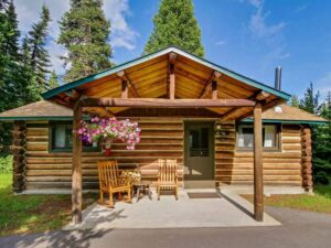 Rustic log cabin with wooden porch, chairs, and flower basket surrounded by evergreen trees under a blue sky in Jenny Lake Lodge