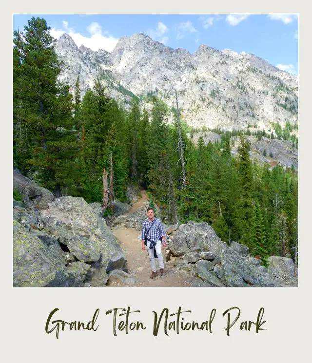 Kevin standing on the trail surrounded by huge rocks and trees behind are mountains in Grand Teton National Park.