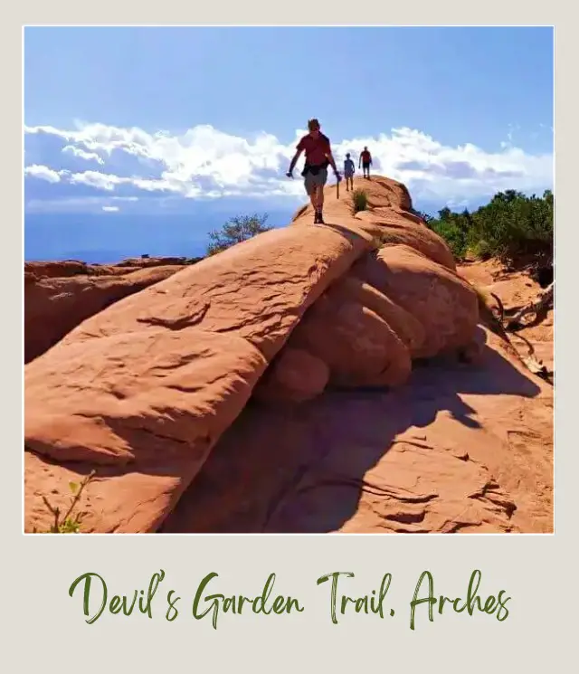 People walking above the huge rock formations surrounded by bushes under the blue skies in Devils Garden in Arches National Park.