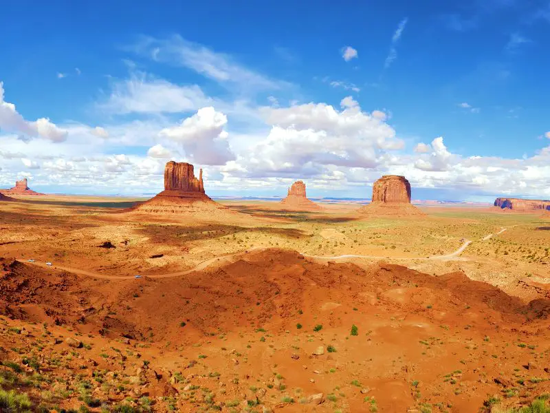 View of different huge rock formations, and below are long roads surrounded by bushes in Monument Valley.