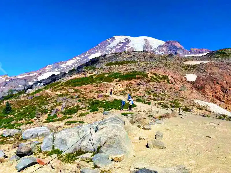 Snow-capped mountains and below are people walking on the rock mountains in Skyline Trail Mt Rainier National.