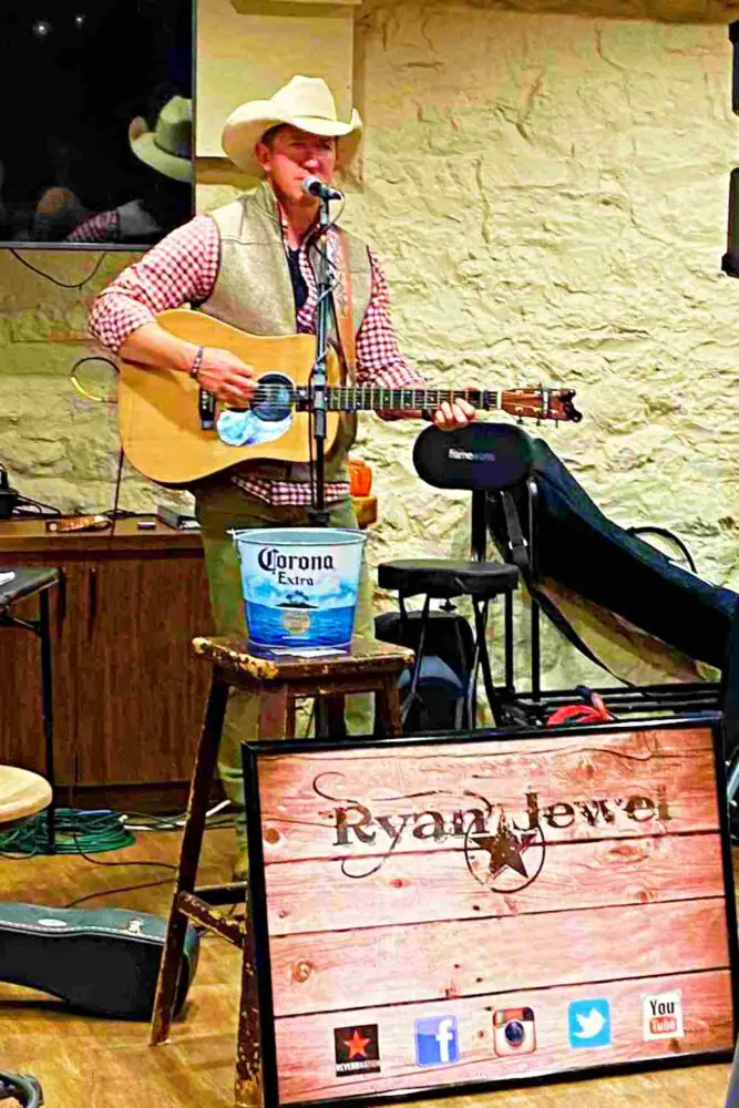 A man in a red checkered long-sleeve singing while holding a guitar surrounded by different things like wires, chairs, and signage that says Ryan Jewel in Shenandoah National Park