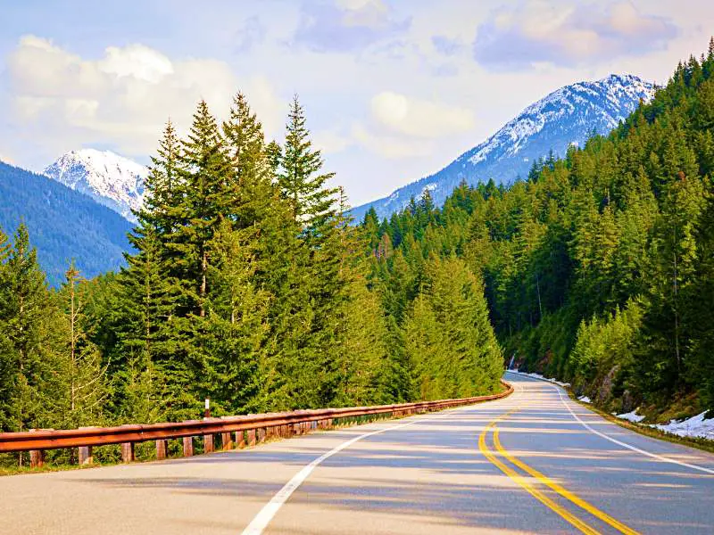 Long and bent road with red railings surrounded by trees and behind are snow-capped mountains in North Cascades National Park