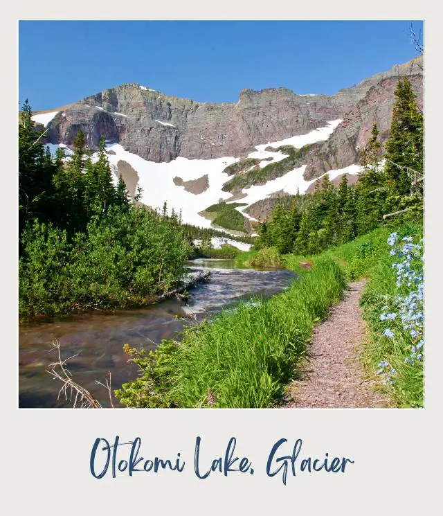 View of a trail beside the lake surrounded by trees and mountains in Otokomi Lake, Glacier National Park.