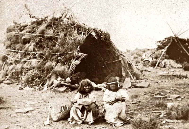 A black and white photo of a two-person sitting in the ground and behind them are wickiup