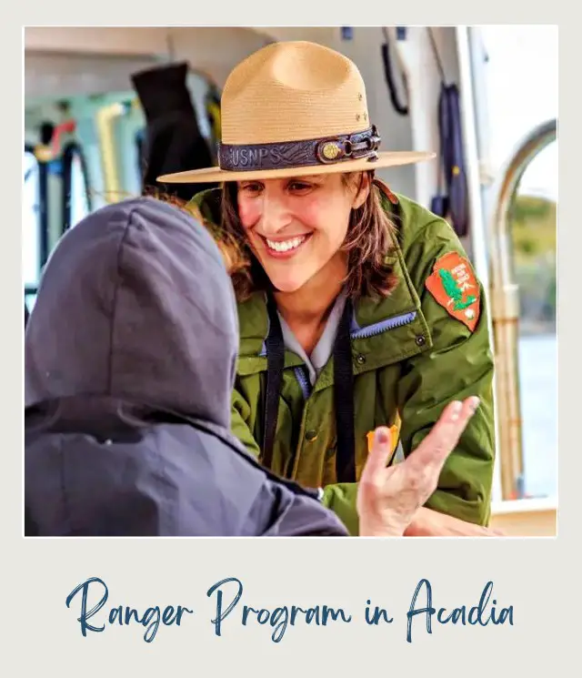 A smiling national park ranger wearing a green Jacket and a brown round cap facing a person with a jacket in Acadia National Park