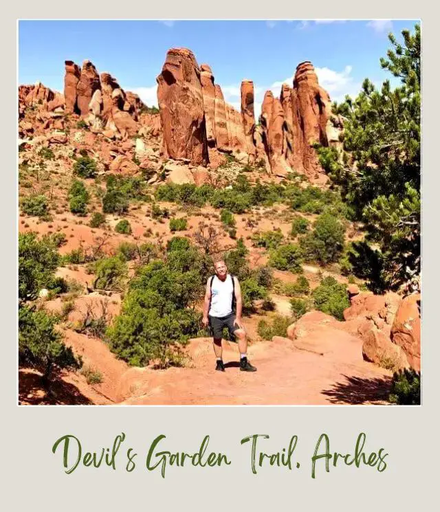 View of James standing on Devils Garden Trail and behind are huge rock formations and bushes.