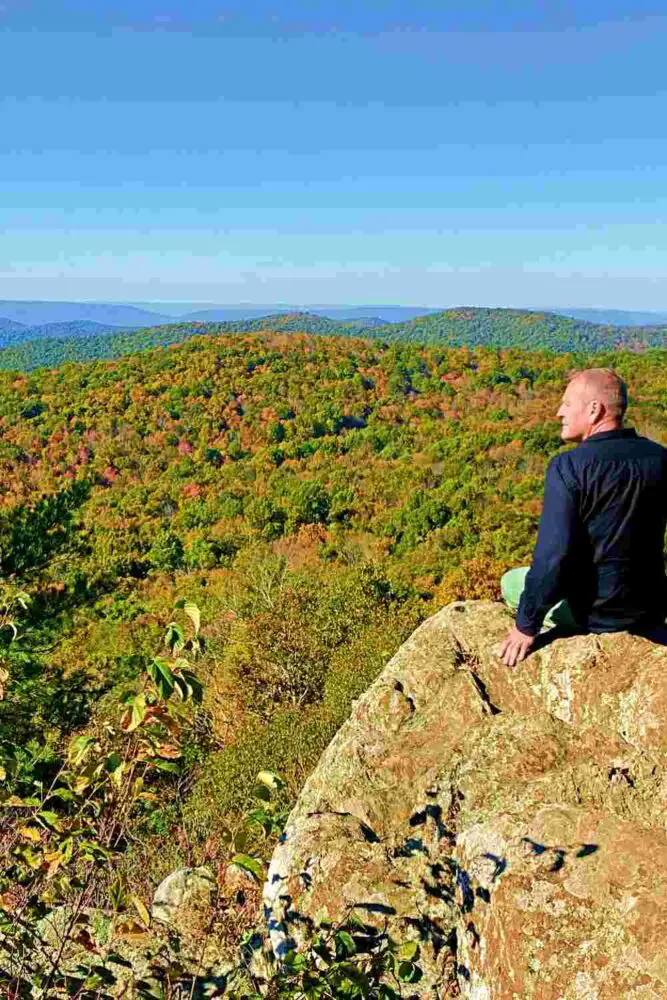 A man is sitting on a huge rock, and in front of Him are trees and mountains in Shenandoah National Park