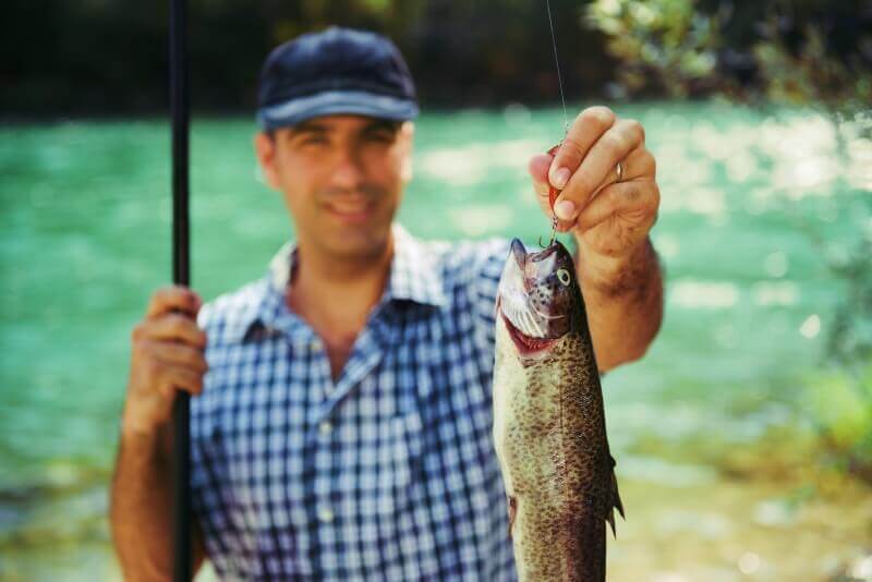 man wearing a cap and holding trout on fishing line with river in background